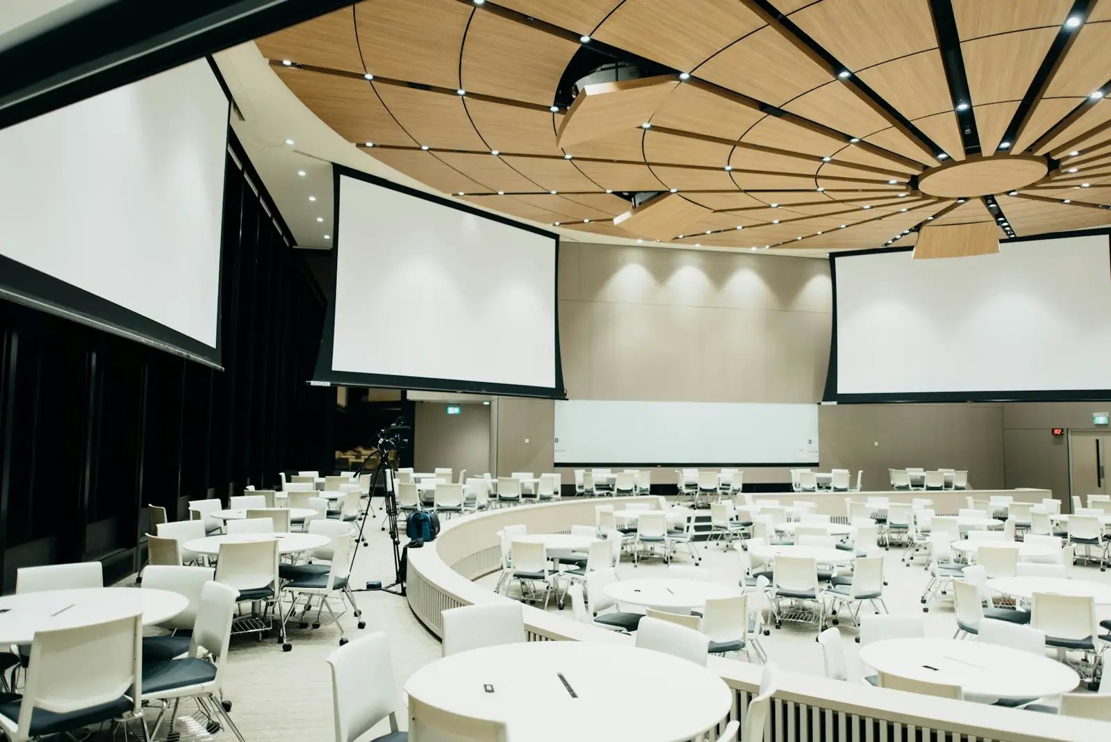 A large conference room with round tables, white chairs, and three projection screens
