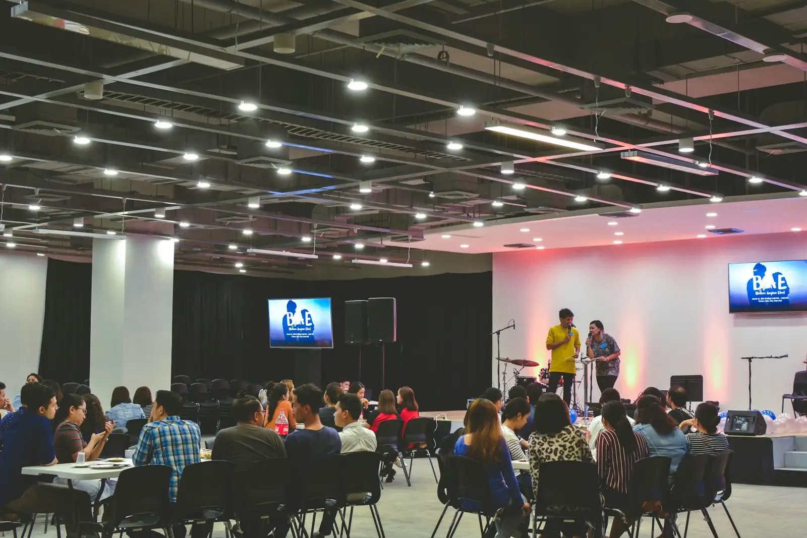 People seated at tables in a large hall watching a presentation on stage